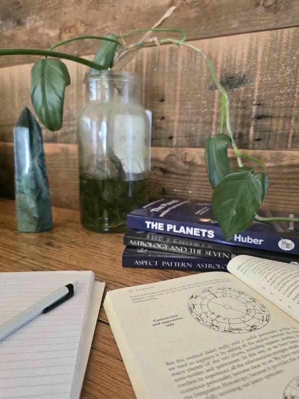 Geburtshoroskop und astrologische Bücher auf einem Tisch. Ein Stift liegt auf dem Notizbuch. Im Hintergrund Bücher zur psychologischen Astrologie der Huber-Schule. Ebenso Edelsteine und Pflanzen.